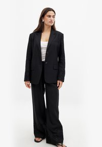 Schwarzer Blazer mit taillierter Passform und zwei Knöpfen, kombiniert mit weit geschnittenen Hosen, die am Saum mit aufwendigen Ösenverzierungen versehen sind.