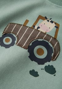 Hellgrüner Stoff mit einem Traktor-Design, das eine Kuh zeigt, umrandet mit braunem Faden, sowie braunen und blauen Akzenten an den Rädern.