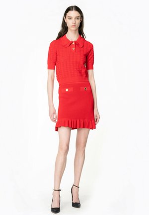 Polo top rosso lavorato a maglia con colletto e maniche corte, abbinato a una mini gonna aderente coordinata con orlo arricciato e dettagli dorati.