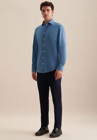 Lichtblauw overhemd met lange mouwen en knopen, met een klassieke kraag, gecombineerd met donkerblauwe broek en zwarte schoenen, staand op een neutrale achtergrond.