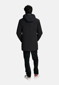 Uomo che indossa un cappotto con cappuccio nero, pantaloni neri e sneakers bianche, in piedi con le mani nelle tasche del cappotto, rivolto di spalle contro uno sfondo semplice.