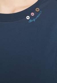 Námořnická modrá trička s kulatým výstřihem, se třemi dekorativními knoflíky a světle modrým vyšitým textem. Měkká látka s hladkým povrchem.