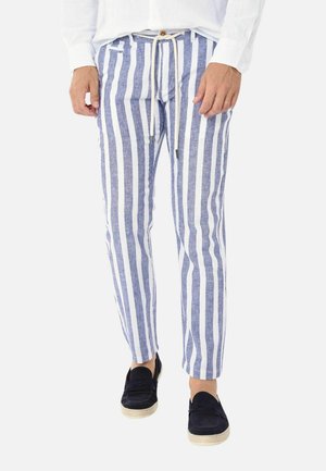Uomo che indossa pantaloni a righe verticali blu e bianchi con coulisse, camicia bianca e mocassini neri, in piedi su sfondo bianco.