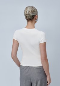 Camiseta blanca ajustada de manga corta, con escote redondo, textura suave y detalles cosidos en la parte de atrás, combinada con pantalones grises.