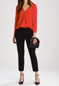 Rød langærmet bluse med V-udskæring, sorte skræddersyede bukser og sort taske. Sorte spidse hæle med ankelremme. Minimalistisk design.