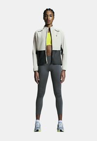 Chaqueta ligera en tonos beige y negro con bloques de color y cuello alto, llevada sobre una blusa amarilla, combinada con leggings grises y zapatillas de running.