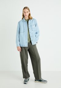 Chemise en denim bleu clair avec boutons sur une chemise verte, associée à un pantalon large olive et des baskets grises avec des accents verts.
