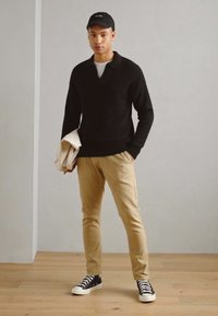 Black knitted sweater, beige tapered pants, black sneakers with white soles, holding a light-colored jacket, wearing a black cap.