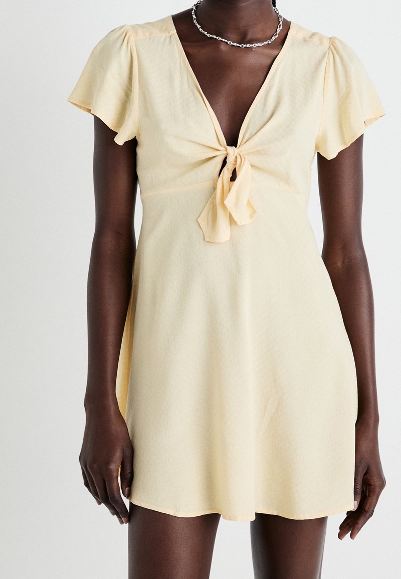 Femme portant une robe jaune pâle à manches courtes avec un lien à l'avant au niveau de la poitrine et un collier en chaîne argenté, sur un fond clair uni.