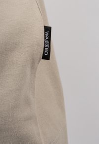 Beige t-shirt med rundad halsringning, tillverkad av mjukt bomull. Har en svart tygetikett med "WASTED"-logotyp på sidan.