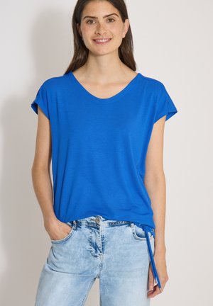 Jeune femme aux longs cheveux bruns portant un t-shirt bleu vif à manches courtes et un jean bleu clair, souriante avec une main dans la poche.