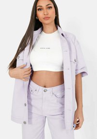 Ljuslila kortärmad tröja och matchande högmidjade jeans, båda med logotryck. Tyget verkar ha en texturerad yta med synliga sömmar och knappar.