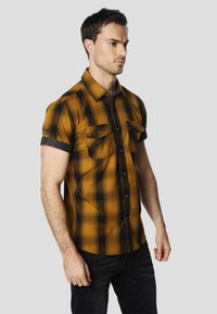 Chemise à manches courtes à carreaux dans des tons ambre et noir, avec deux poches poitrine, fermeture boutonnée à l'avant, et col ainsi que poignets contrastants foncés.