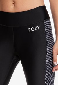 Detailní záběr na černé legíny s logem "ROXY" na levém boku a bílým opakujícím se textovým vzorem na pravém bočním panelu.