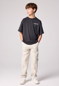 Fekete pamut póló fehér "ALLSAINTS" logóval, krémszínű nadrággal és fehér tornacipővel párosítva, laza szabással.