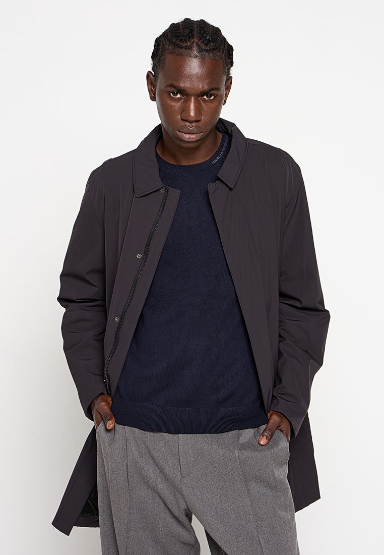 Ciemnobrązowy lekki płaszcz z kołnierzykiem, zapięciem na guziki i długimi rękawami, noszony na granatowym swetrze i szarych spodniach.