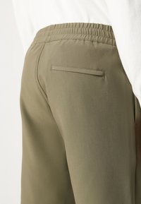 Olive green pants with an elastic waistband, featuring a single back pocket and smooth texture, shown from a side angle.