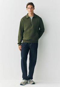 Olivgrüner Pullover mit halbem Reißverschluss, Kängurutasche und gerippten Bündchen, kombiniert mit dunkelblauen Jeans und grauen Sportschuhen.