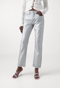Jeans plateados metálicos con corte recto, cinco bolsillos y detalles de costura sutiles, combinados con sandalias de tiras moradas claras.