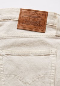 Vaaleanväriset farkut, joissa on nahkainen merkki vyötäröllä. Merkissä lukee "Levi Strauss & Co." ja muita tekstidetaljeja, näkyvissä tikatut taskut.