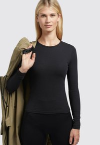 Maglietta nera a maniche lunghe con una texture a coste, abbinata a una giacca beige. La modella tiene la giacca con una tracolla nera etichettata "KHLL."