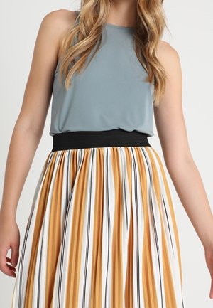 Nainen, jolla on päällään hihaton sininen toppi, joka on tungettu korkeavyötäröisen, liehuvan hameen sisään, jossa on pystysuuntaiset sinapin, valkoiset ja mustat raidat.