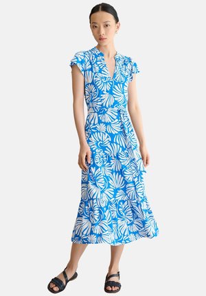 Mujer vestida con un vestido midi azul y blanco con estampado floral, mangas cortas y sandalias negras abiertas en los dedos, de pie frente a un fondo liso.