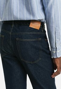 Jeans de mezclilla azul oscuro con costuras amarillas, que presentan un parche de cuero marrón claro con el logo de GANT en la parte posterior, combinados con una camisa de rayas azul clara.