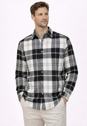 Uomo che indossa una camicia a quadri nera, bianca e grigia abbottonata, con pantaloni beige, mani in tasca, che guarda di lato con un leggero sorriso.