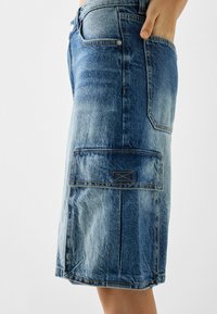 Jupe en denim bleu clair, longueur au genou, avec poches latérales et détails de couture. Le tissu présente une texture légèrement usée.