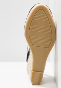 Semelle en espadrille avec un dessus en jute beige texturé et des sangles en coton noir. Semelle extérieure en caoutchouc plate et flexible avec un motif strié et une étiquette "Fabriqué en Espagne".