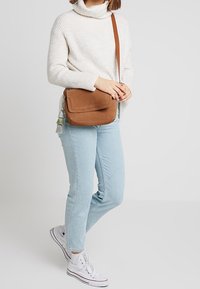 Ruskea mokkagootti olkalaukku, jossa on läppä suljenta ja koristeellinen tupsu, käytetään valkoisen neulotun neuleen ja vaaleansiniset farkut päällä.