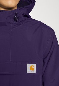 Lilla hættejakke med lynlås, justerbare snore og et hvidt mærke med et orange logo, lavet af letvægtsmateriale.
