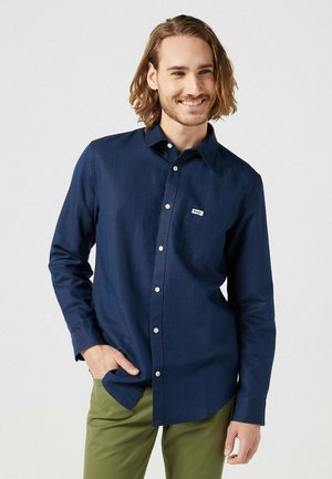 Camicia azzurra scuro con maniche lunghe, con bottoni in un tessuto liscio e intrecciato, dotata di una piccola tasca frontale e bottoni bianchi, indossata con pantaloni verdi.