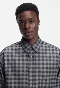 Harmaa-musta ruudullinen kauluksellinen nappipaita, jossa on rintatasku ja näkyvät napit. Pehmeä kangas ja ryhdikäs muoto.