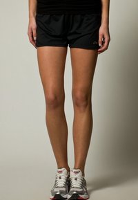 Shorts de sport noirs en tissu léger, avec une taille élastique. Portés avec des chaussures de sport grises et blanches, mettant en avant des lacets et un rembourrage.