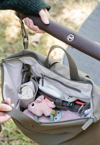 Kädet pitävät vaunuista kiinni, joissa on avoin vaippalaukku, joka sisältää vauvan tarvikkeita, vaaleanpunaisen pehmolelun, tutin ja taiteltuja vaatteita.