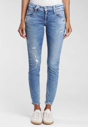 Frau trägt hellblaue, ausgewaschene Skinny-Jeans mit rohem Saum und weißen Sneakers, steht vor einem schlichten Hintergrund.