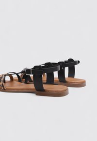 Sandalias planas de estampado de leopardo con tiras negras, hebillas ajustables y suelas marrones. Presentan un empeine texturizado y diseño de punta abierta.