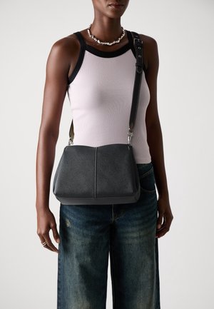 Femme portant un haut blanc sans manches et un jean bleu, portant un sac bandoulière en cuir noir avec une large sangle ajustable.