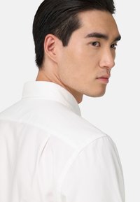 Camicia button-up bianca in cotone con colletto a punta, mostra cuciture ordinate e una texture liscia; la testa del modello è leggermente inclinata a sinistra, mettendo in risalto il profilo.