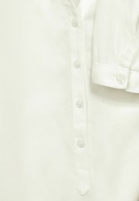 Camicia bianca con colletto classico, pattina con sei bottoni e maniche arrotolate. Realizzata in un tessuto leggero con una texture liscia.