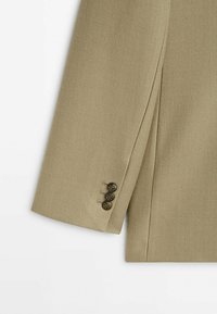 Manche de costume beige avec trois boutons décoratifs marron et détails de coutures visibles sur un fond en tissu texturé.