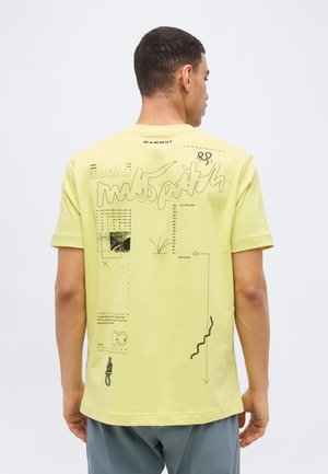 Άνδρας που φοράει κίτρινη μπλούζα Mammut με αφηρημένα τεχνικά και αναρριχητικά γραφικά στην πλάτη, γυρισμένος με την πλάτη στην κάμερα.