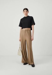 Top negru cropped, pantaloni bej plisati cu croiala largă, textură netedă, design minimalist. Modelul ține o geantă mică bej și poartă pantofi negri comozi.