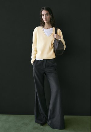 Vrouw met een gele V-hals trui over een wit shirt, zwarte broek met wijde pijpen, een donkerbruine handtas vasthoudend, staand op een groene vloer.