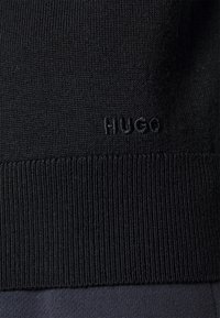 Tissu en tricot noir avec texture côtelée et bande. Présente un logo « HUGO » ton sur ton brodé sur le côté. Fini lisse, design ajusté.