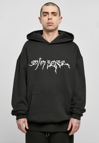 Mann mit kurzen Haaren, der einen schwarzen Kapuzenpullover mit weißem, stilisiertem Text "ontm sense" trägt und vor einem einfarbigen grauen Hintergrund steht.