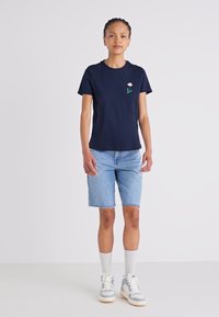 T-shirt de algodão azul-marinho com um pequeno bordado floral; combinado com calções de ganga claros e sapatilhas altas cinzas, com meias brancas.