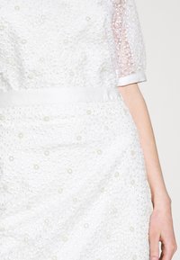 Robe blanche en tissu texturé, avec des motifs floraux brodés et des accents en perles. Manches courtes et transparentes avec une ceinture en satin.